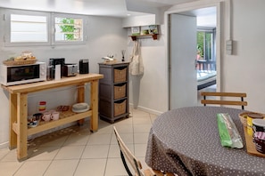 A compact kitchen with a microwave, toaster, and coffee maker on a wooden table. There is a wicker basket and a small shelf with books and a lamp. A chair is placed at a round table with a polka dot tablecloth.
