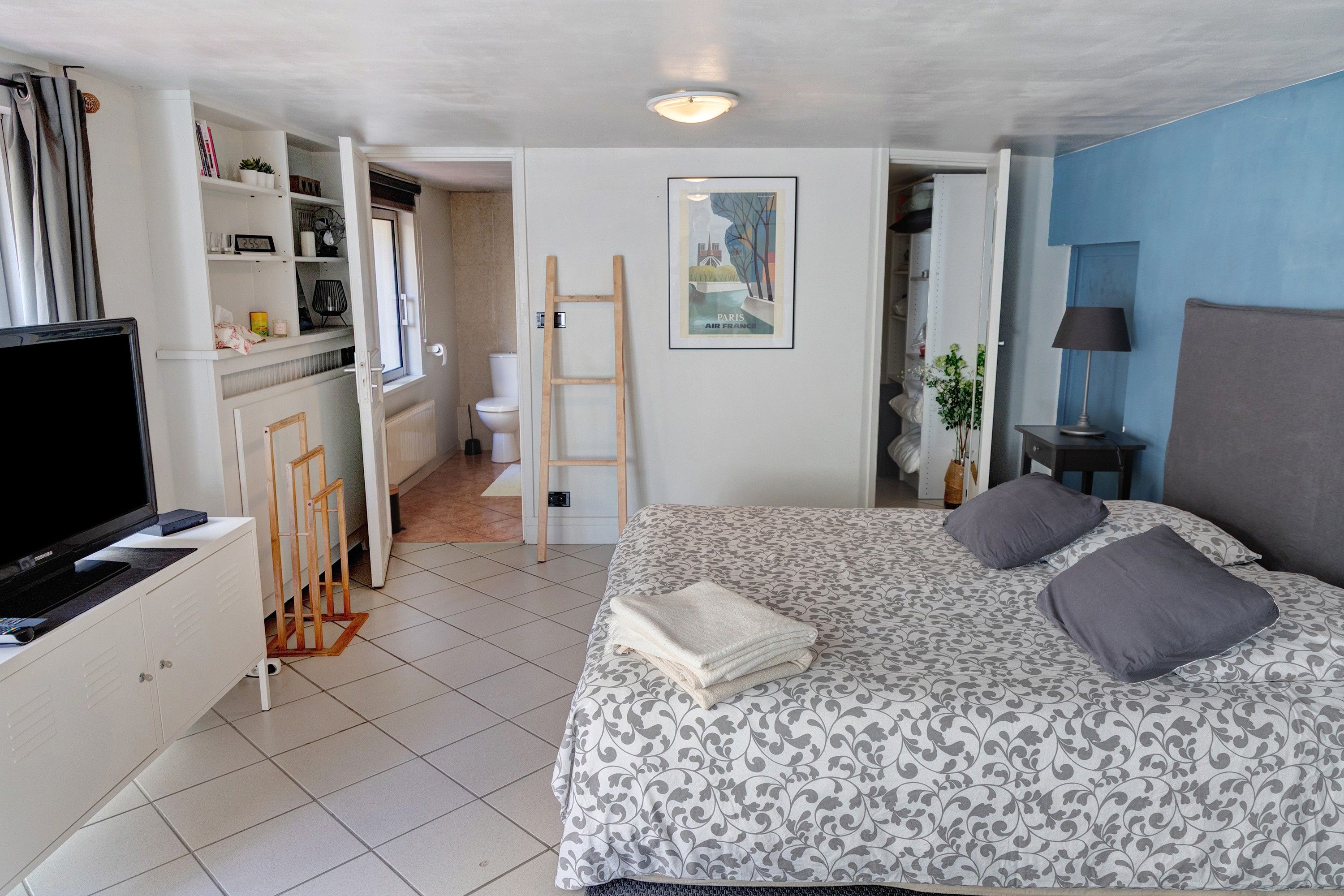 Quarto com cama, televisão, guarda-roupa, banheiro e um quadro na parede.