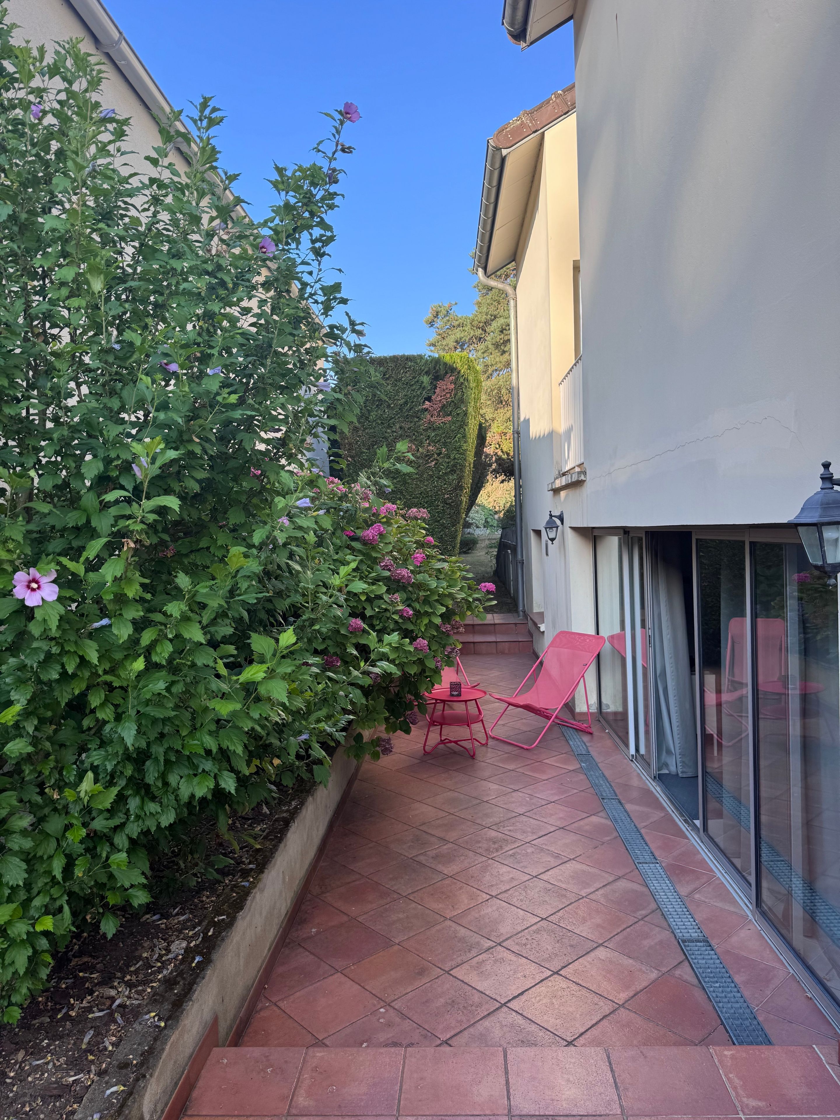 A patio with a red chair and a small table, a hedge with purple flowers, and a building with a white exterior.