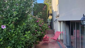 A patio with a red chair and a small table, a hedge with purple flowers, and a building with a white exterior.
