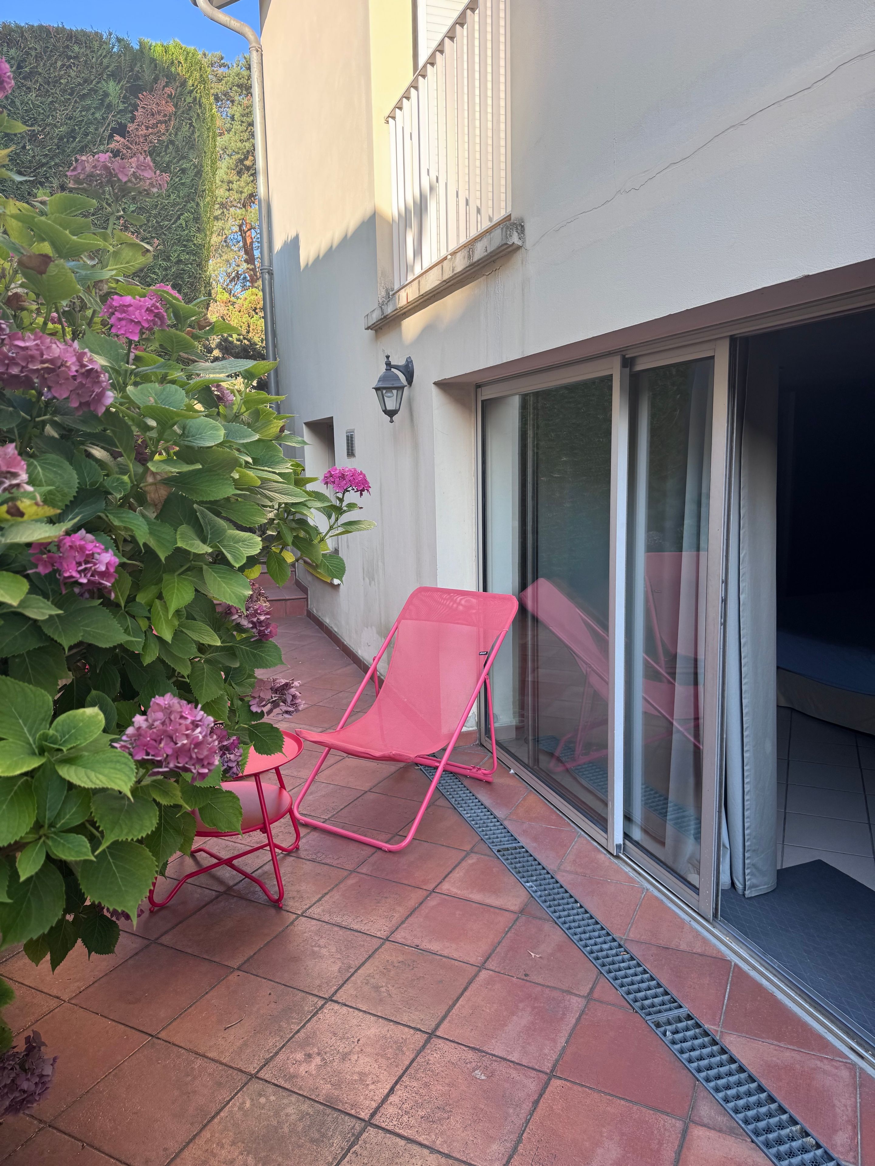 Een terras met een rode stoel, een potplant met roze bloemen en een schuifdeur.