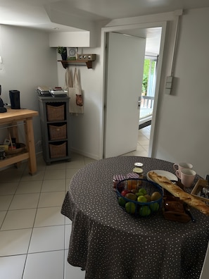 Une cuisine avec une table ronde recouverte d’une nappe à pois, un panier de fruits et une baguette.