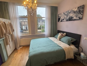 Ein Schlafzimmer mit einem Bett, einer Chandelüster, einem Gemälde und Blick auf Gebäude draußen.