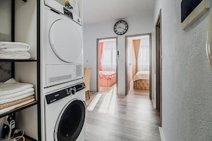 Un área de lavandería con lavadora y secadora, un reloj en la pared y un pasillo que conduce a un dormitorio con cama y cortinas.