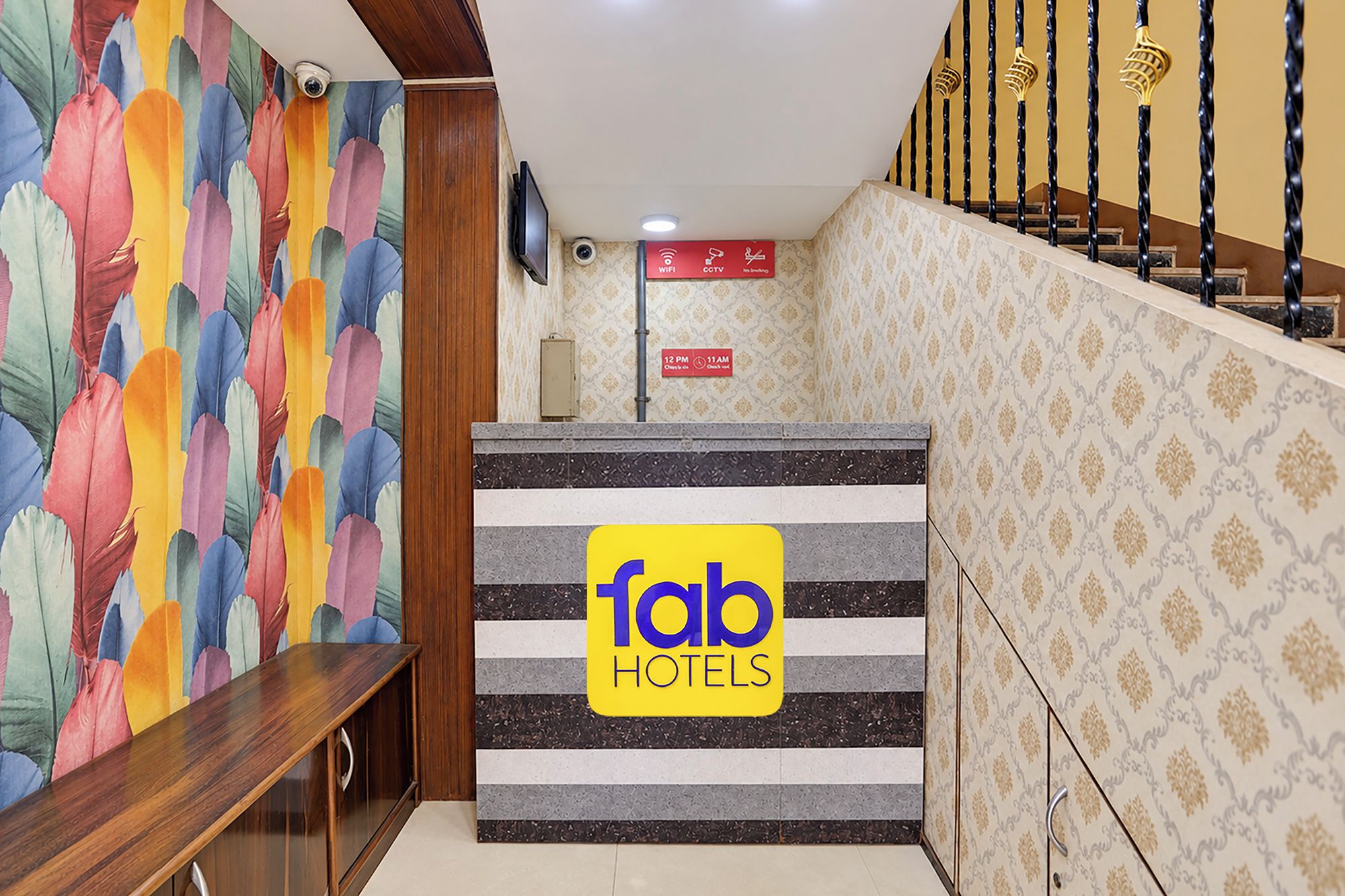 Uma área de recepção de hotel com parede decorada com padrão de penas coloridas, balcão de atendimento com a placa 'fab hotels', uma câmera de vigilância e escada com parede estampada.