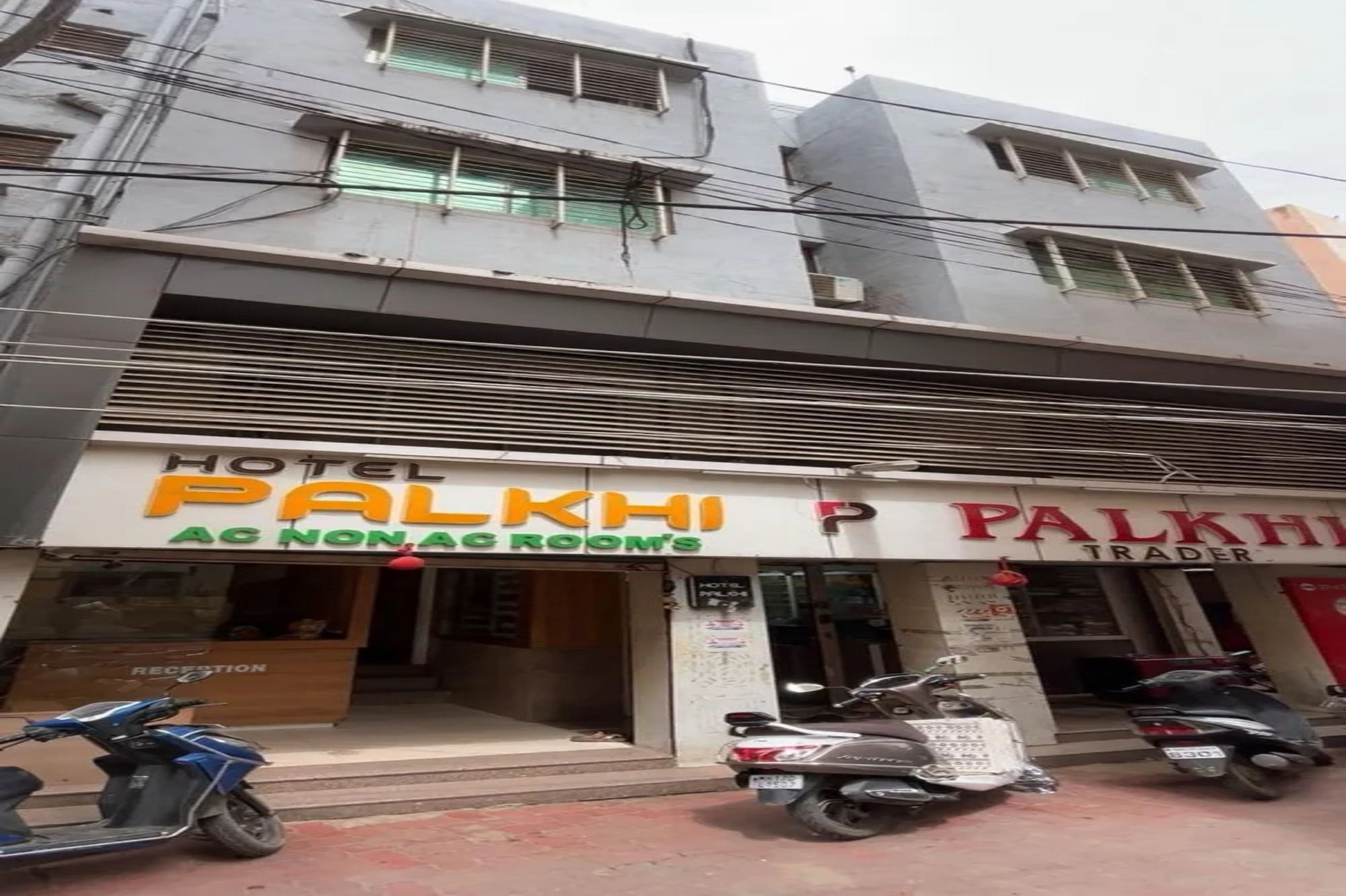 Hotel Palkhi, con un cartel que indica 'Habitación con aire acondicionado/sin aire acondicionado' y una sección 'Trader'. Hay scooters estacionados al frente.