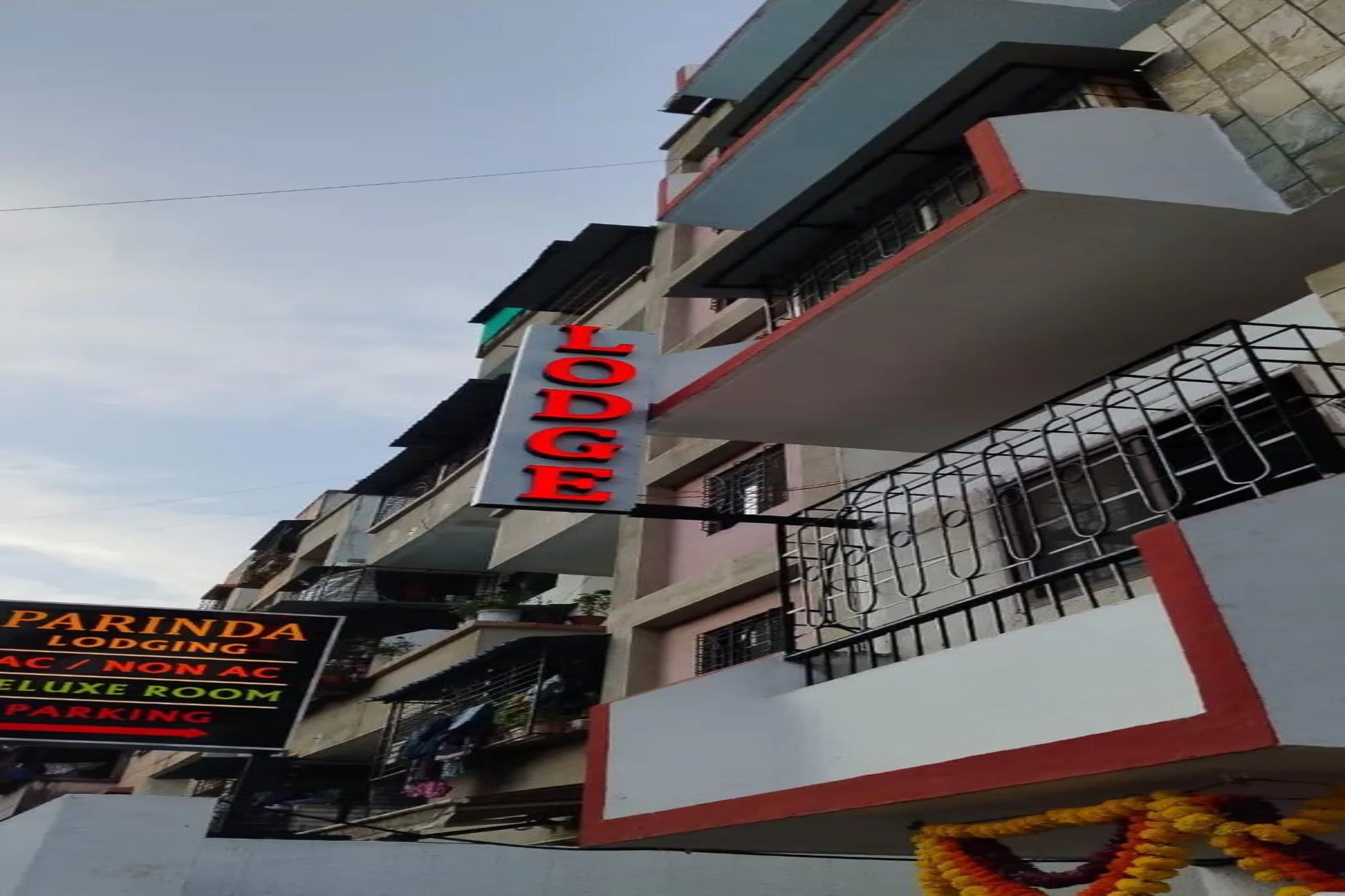 Un edificio de varios pisos con un letrero destacado que dice 'Lodge' y un cartel anunciando 'Parinda Lodging', indicando 'Aire acondicionado / Sin aire acondicionado' y 'Habitación Deluxe', con 'Estacionamiento' disponible.