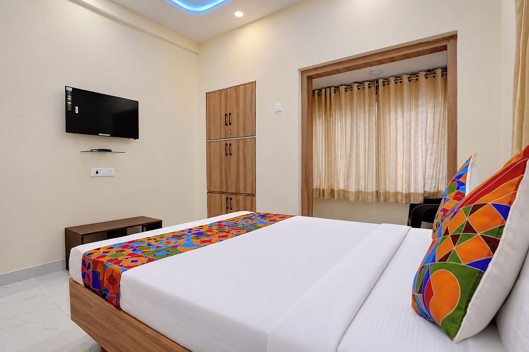 A hotel room with a bed, a television mounted on the wall, a wardrobe, and a window with curtains.