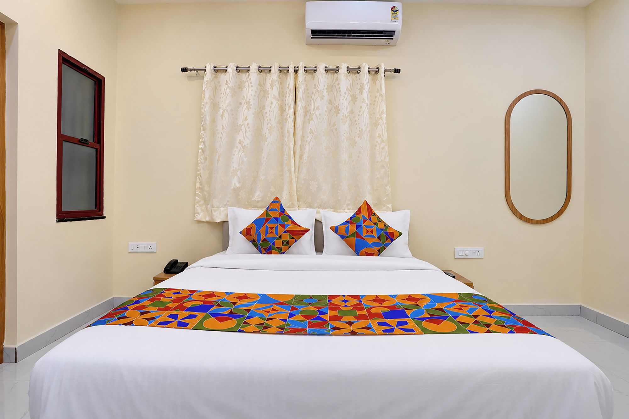 Een hotelkamer met een bed, een raam met gordijnen, een airconditioningapparaat, een wandspiegel en een telefoon op het nachtkastje.
