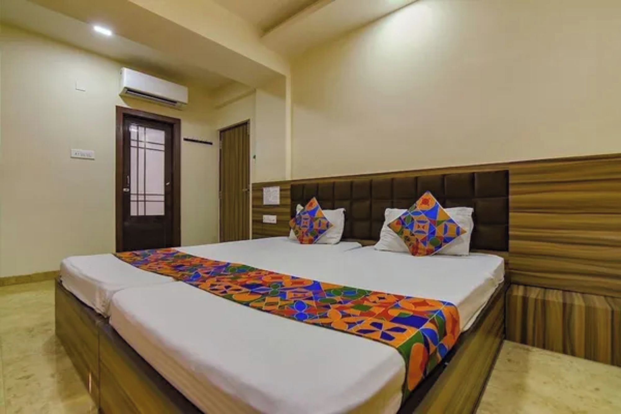 A hotel room with a bed, a wooden headboard, a colorful bedspread, and a door with a window.