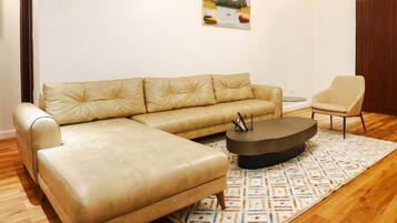 A beige leather sectional sofa, a round coffee table, and a single armchair in a living room with wooden flooring and a patterned rug.