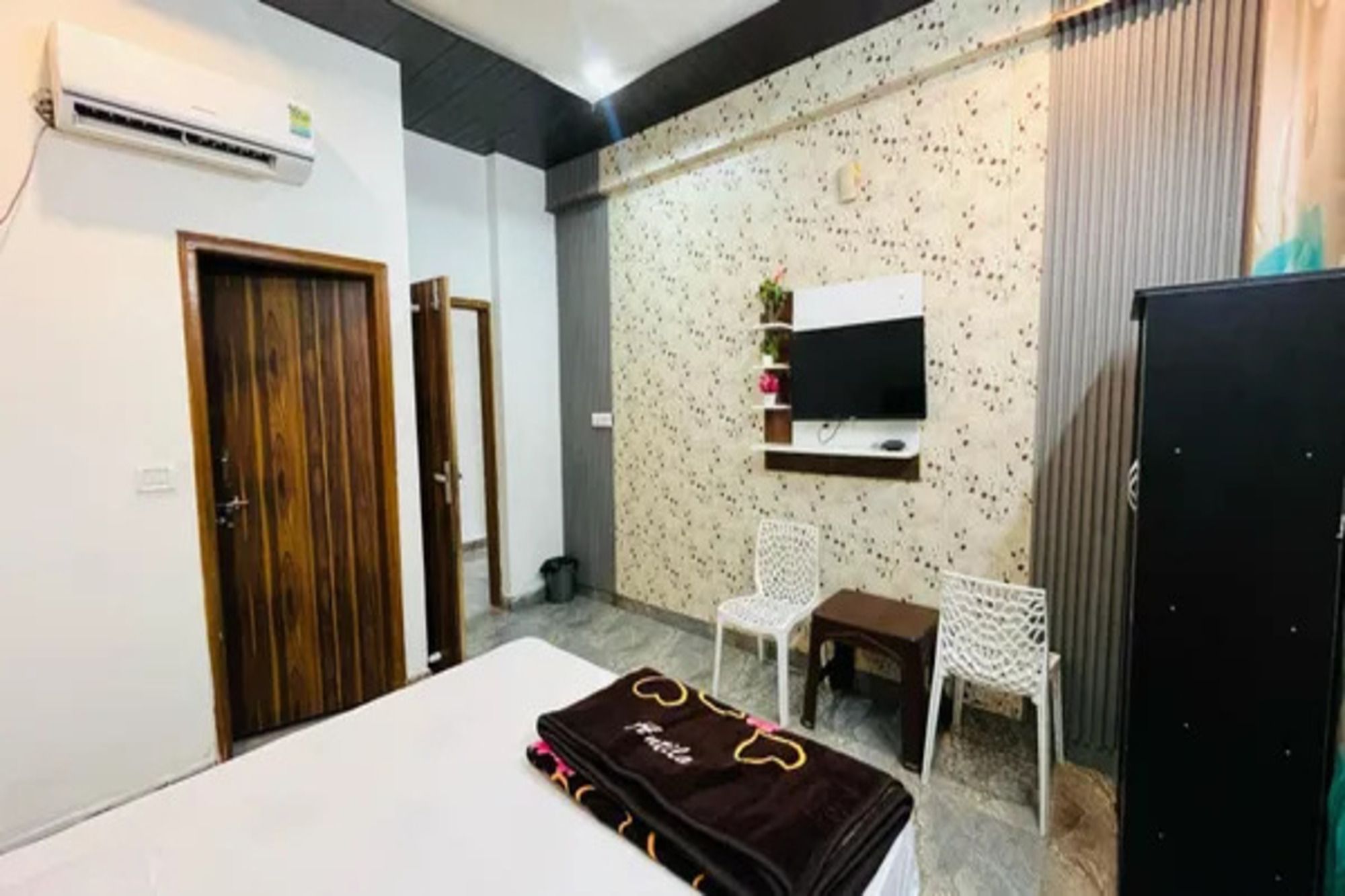Un dormitorio con cama, televisor montado en la pared, una mesita, dos sillas y un armario.