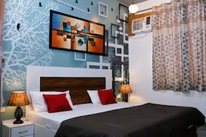 Una habitació d'hotel amb un llit, dues làmpades de nit, una paret amb un quadre geomètric i una finestra amb cortines.