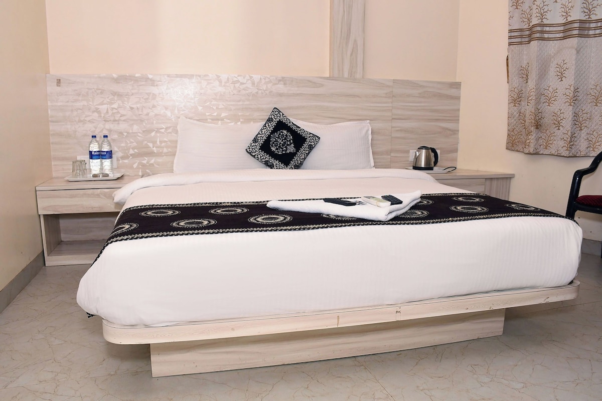 Una habitación de hotel con cama, mesita de noche con una botella de agua, tetera y una silla.