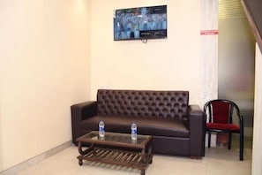 Un hall con un sofá marrón oscuro, una mesa de centro de vidrio con botellas de agua, un televisor montado en la pared y una silla roja.