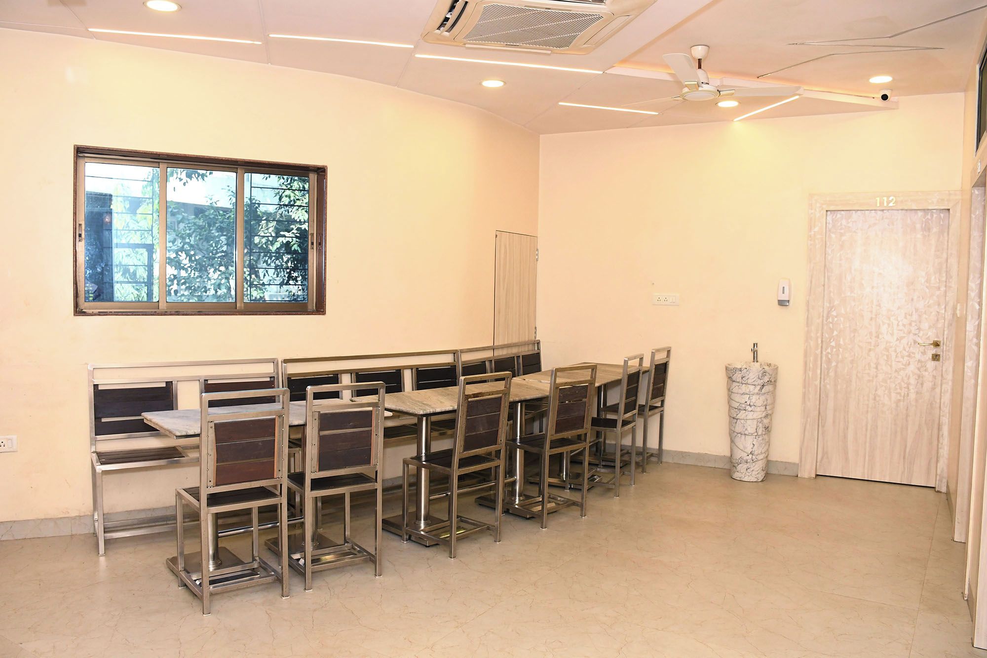 Un salón de conferencias con una larga mesa y sillas, una ventana con vistas a árboles, un ventilador de techo y una puerta.