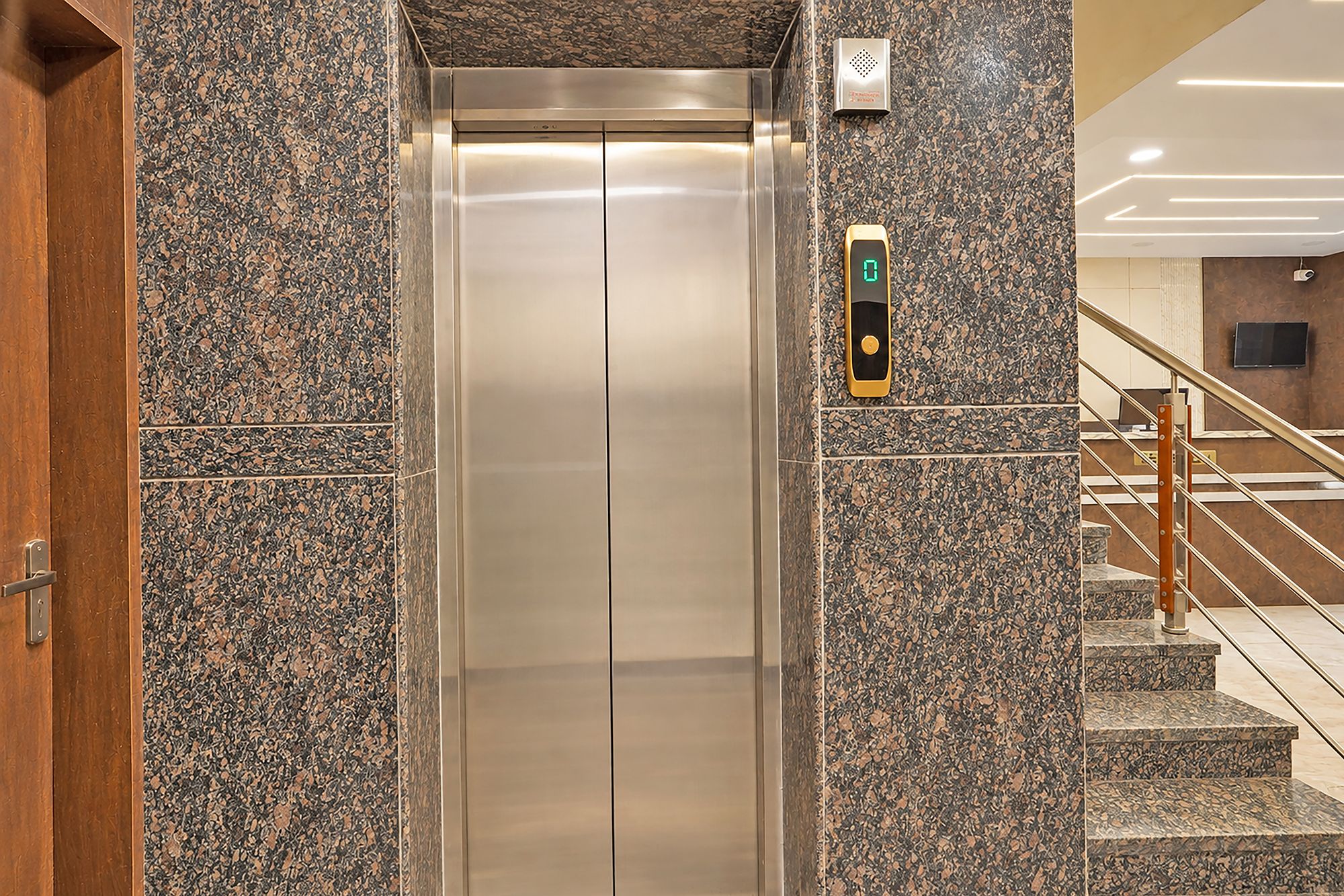 Un ascensor con pantalla digital, flanqueado por paredes de granito y una puerta de madera.