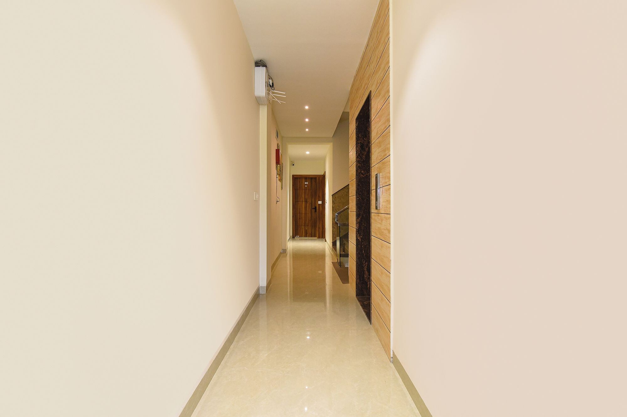 Un pasillo estrecho con una puerta de madera al final, un equipo de aire acondicionado en la pared y un pasamanos a la derecha.