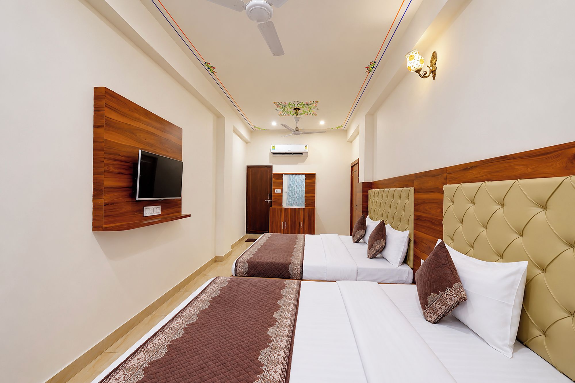 Habitación de hotel con una televisión de pantalla plana montada en la pared, un ventilador de techo, una lámpara decorativa y una cama con cabecera y almohadas.