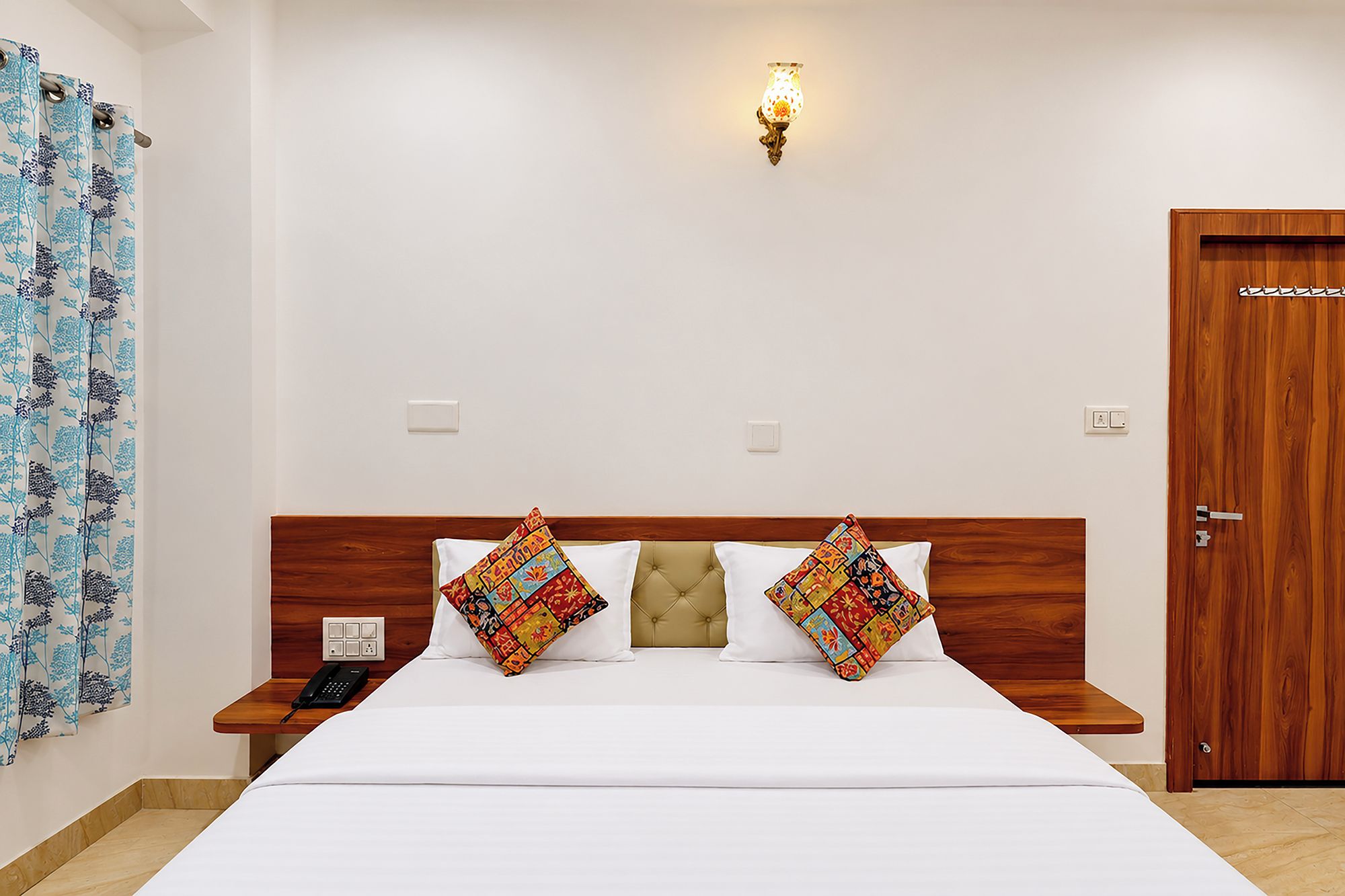 Quarto de hotel com cama, duas mesas de cabeceira, abajur, porta e cortina estampada.