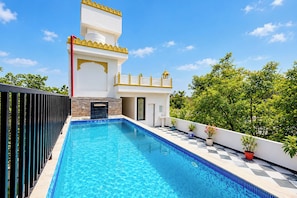 Un área de piscina moderna con una piscina azul cristalina, un edificio blanco con detalles amarillos y abundante vegetación al fondo.