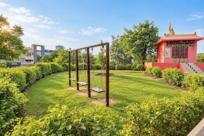 A well-maintained garden with a swing set, a small red structure with steps, and a building with 'INFINITE' signage in the background.