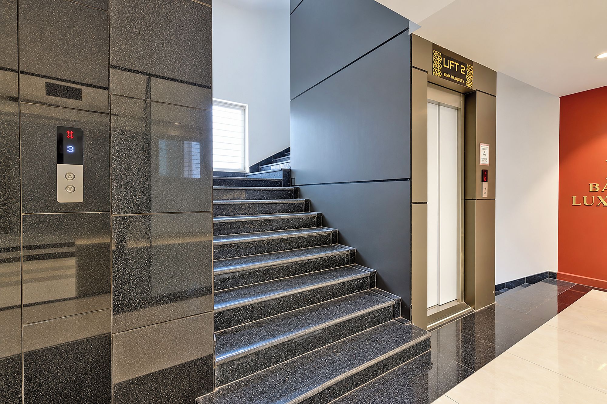 Un ascenseur moderne doté d’un écran numérique et de boutons, un escalier avec des marches à motifs sombres et un mur rouge orné de l’inscription « BISMA BANQUETS ».