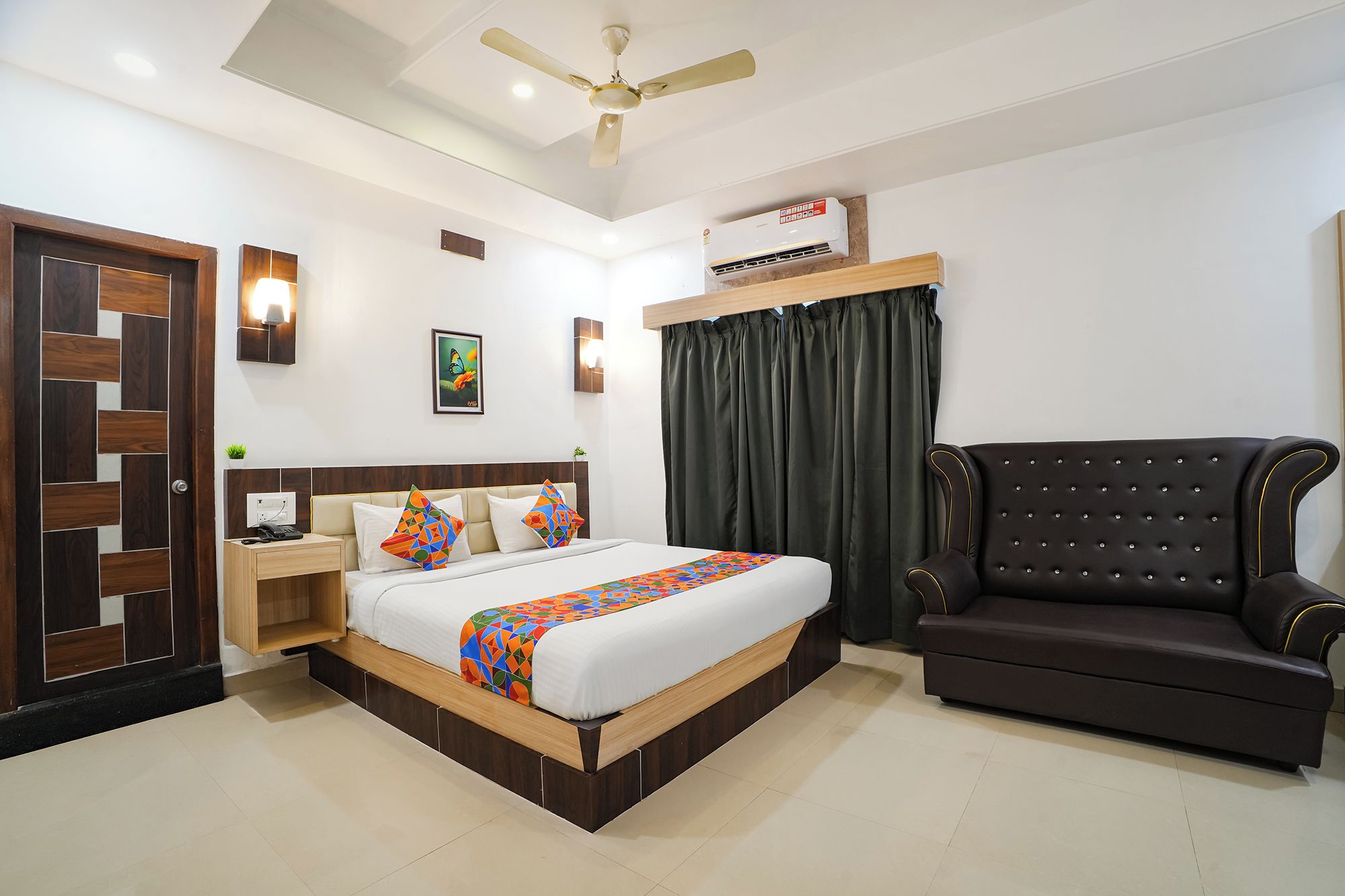 A hotel room with a bed, bedside table, lamp, air conditioning unit, and a dark-colored sofa.