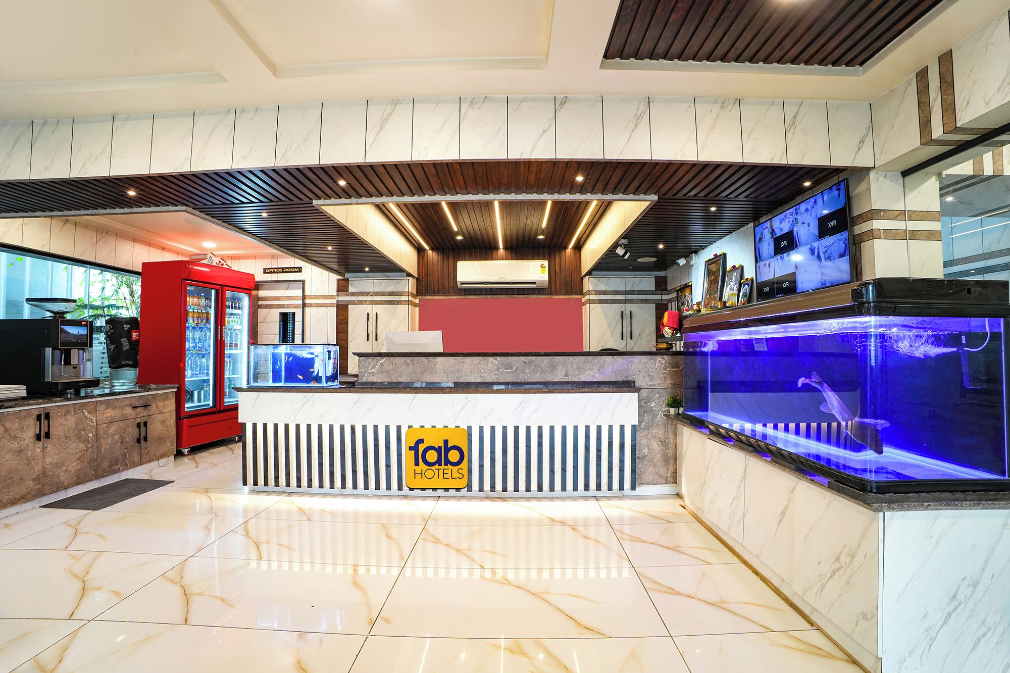 Zona de recepció d'un hotel amb sòl de marbre, una taula de recepció, un refrigerador vermell, un aqüari i una pantalla digital que mostra informació sobre les habitacions.