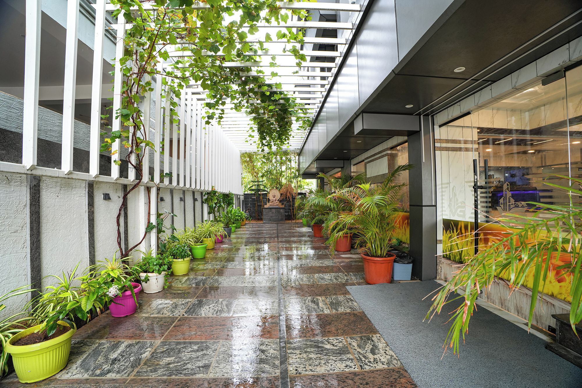 A covered outdoor corridor with potted plants, a wet floor, and a staircase leading to an upper level.