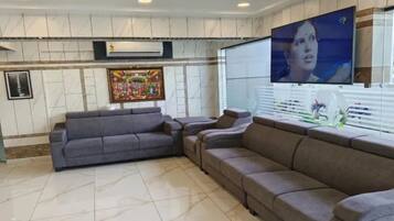 Een woonkamer met een hoekbank, een flatscreen-televisie aan de wand en een groot raam met zonwering.