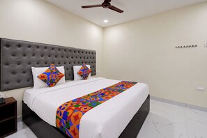 Un dormitorio con cabecera capitonada, cama con ropa de cama blanca, dos almohadas de colores, ventilador de techo y piso de baldosas.