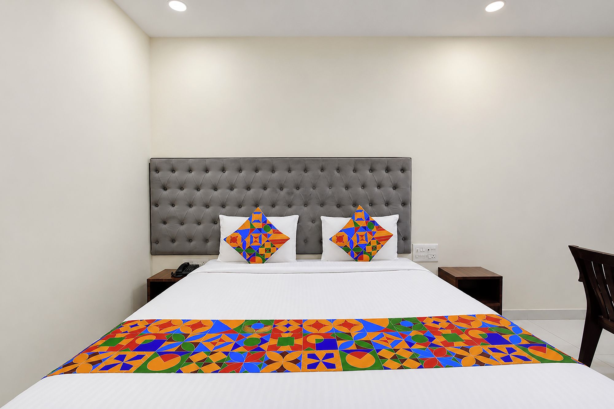 A hotel room with a tufted headboard, a large bed with a colorful geometric-patterned bedspread, two pillows with geometric designs, and a nightstand with a phone.