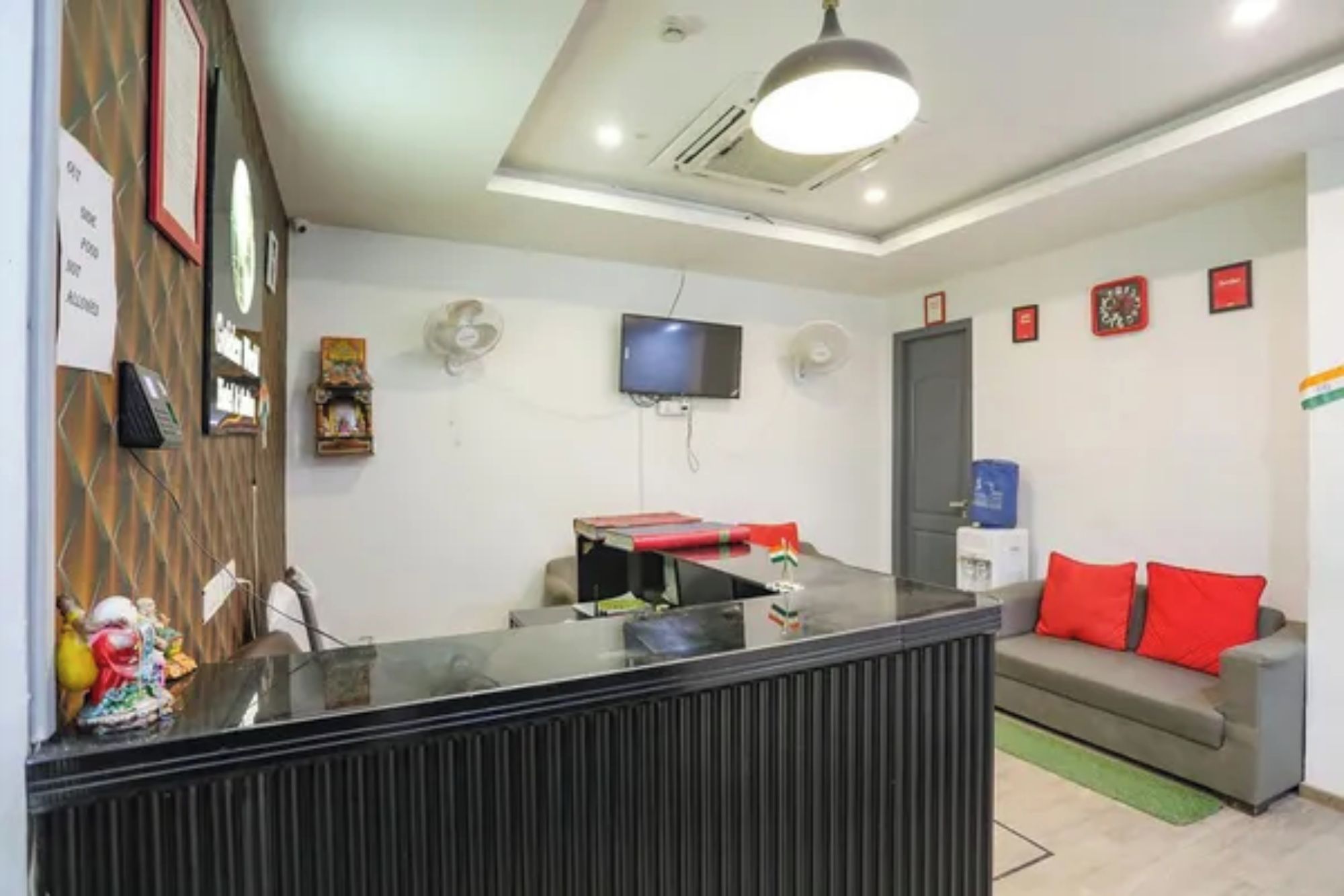 Un área de recepción con mostrador, un televisor montado en la pared, un sofá con cojines rojos y una bandera en la pared.