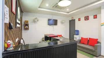 Un área de recepción con mostrador, un televisor montado en la pared, un sofá con cojines rojos y una bandera en la pared.