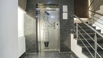 Un ascensor con una persona sosteniendo un teléfono, una escalera con barandillas de metal y una pared con una placa.
