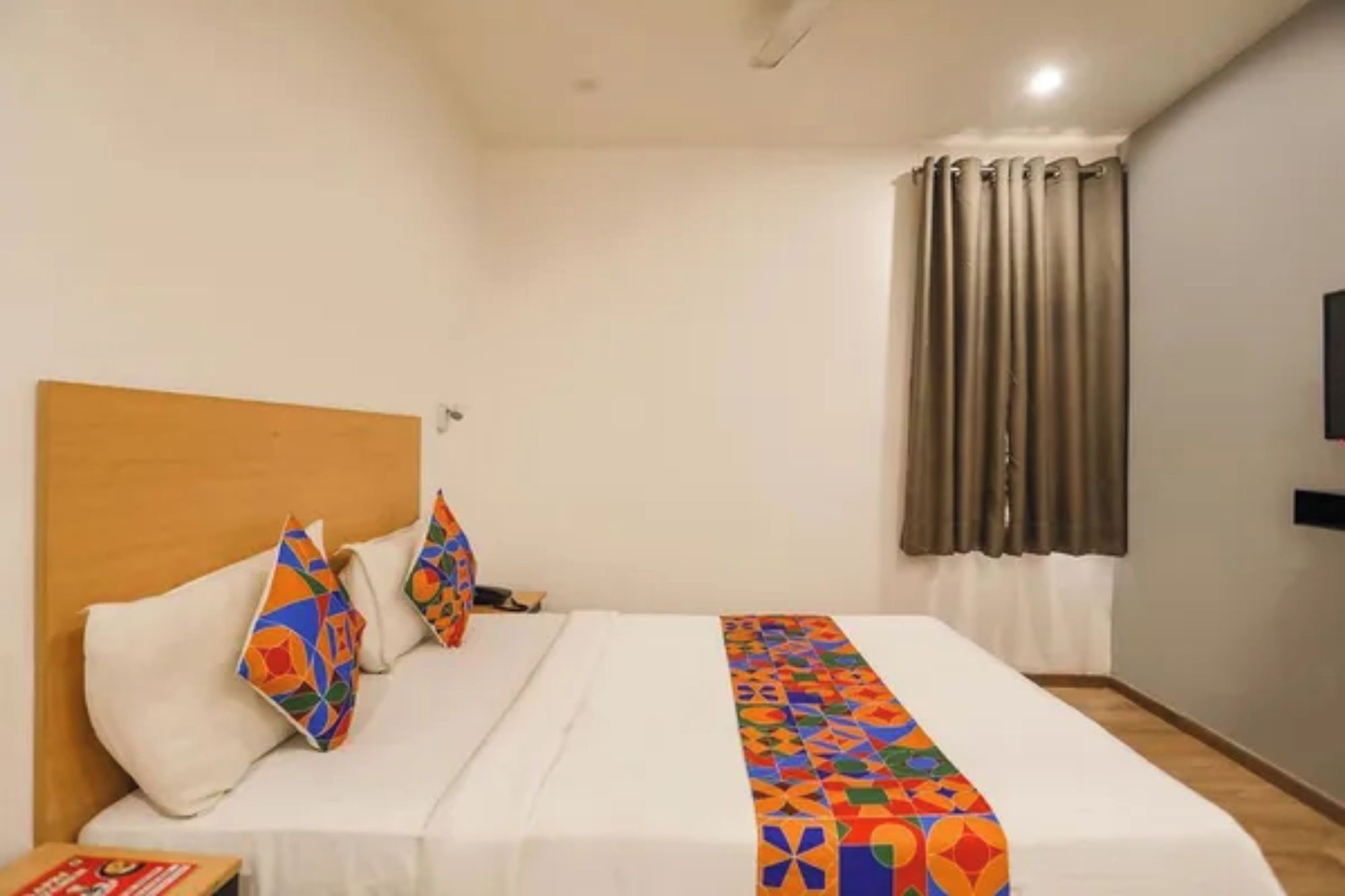 A hotel room with a bed, pillows, a blanket, a wooden headboard, a window with curtains, and a flat-screen TV on the wall.