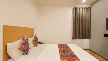 A hotel room with a bed, pillows, a blanket, a wooden headboard, a window with curtains, and a flat-screen TV on the wall.