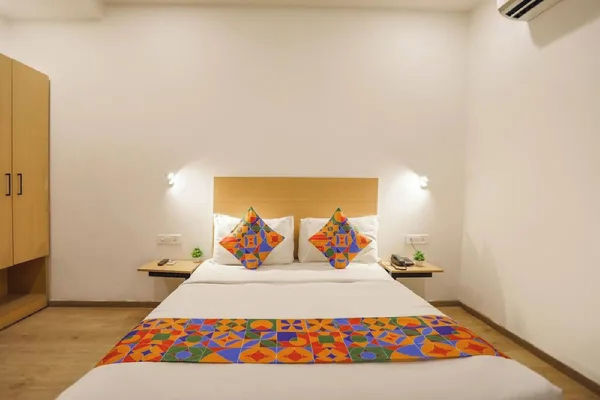 Una habitación de hotel con una cama, dos mesitas de noche, un tablero auxiliar, cabecera de madera y un aire acondicionado instalado en la pared.