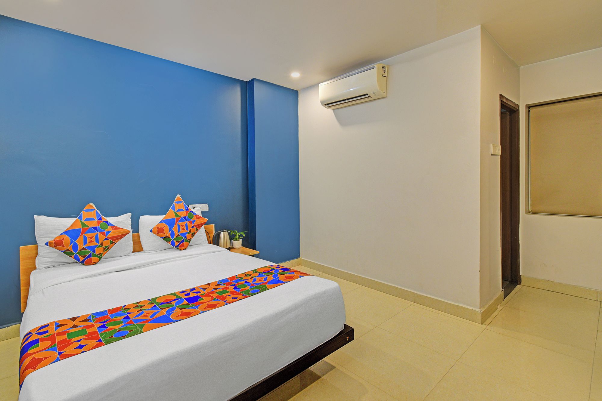 A hotel room with a bed, a nightstand, an air conditioning unit, and a door.