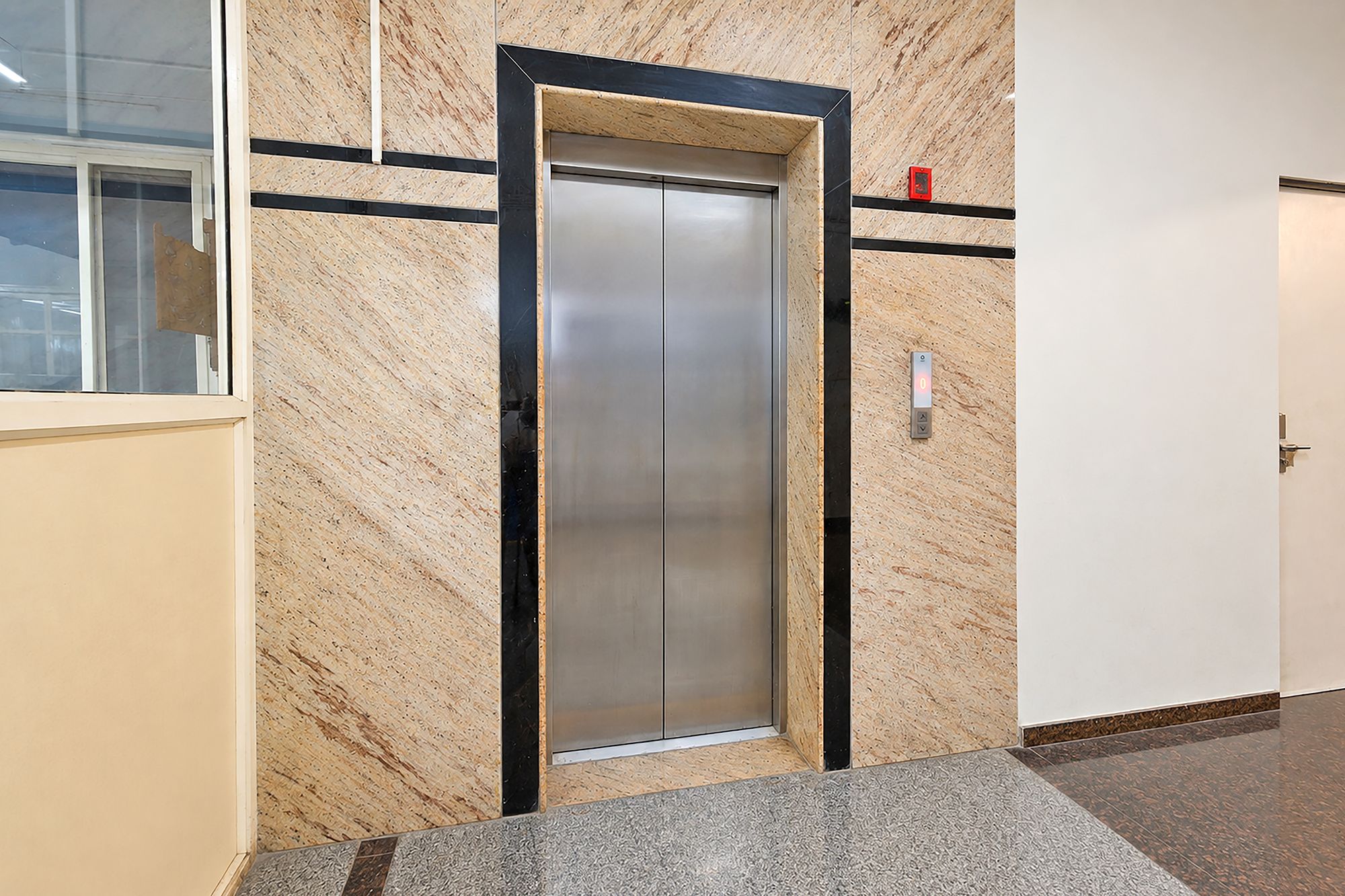 An elevator in a hallway with a marble wall and a tiled floor.