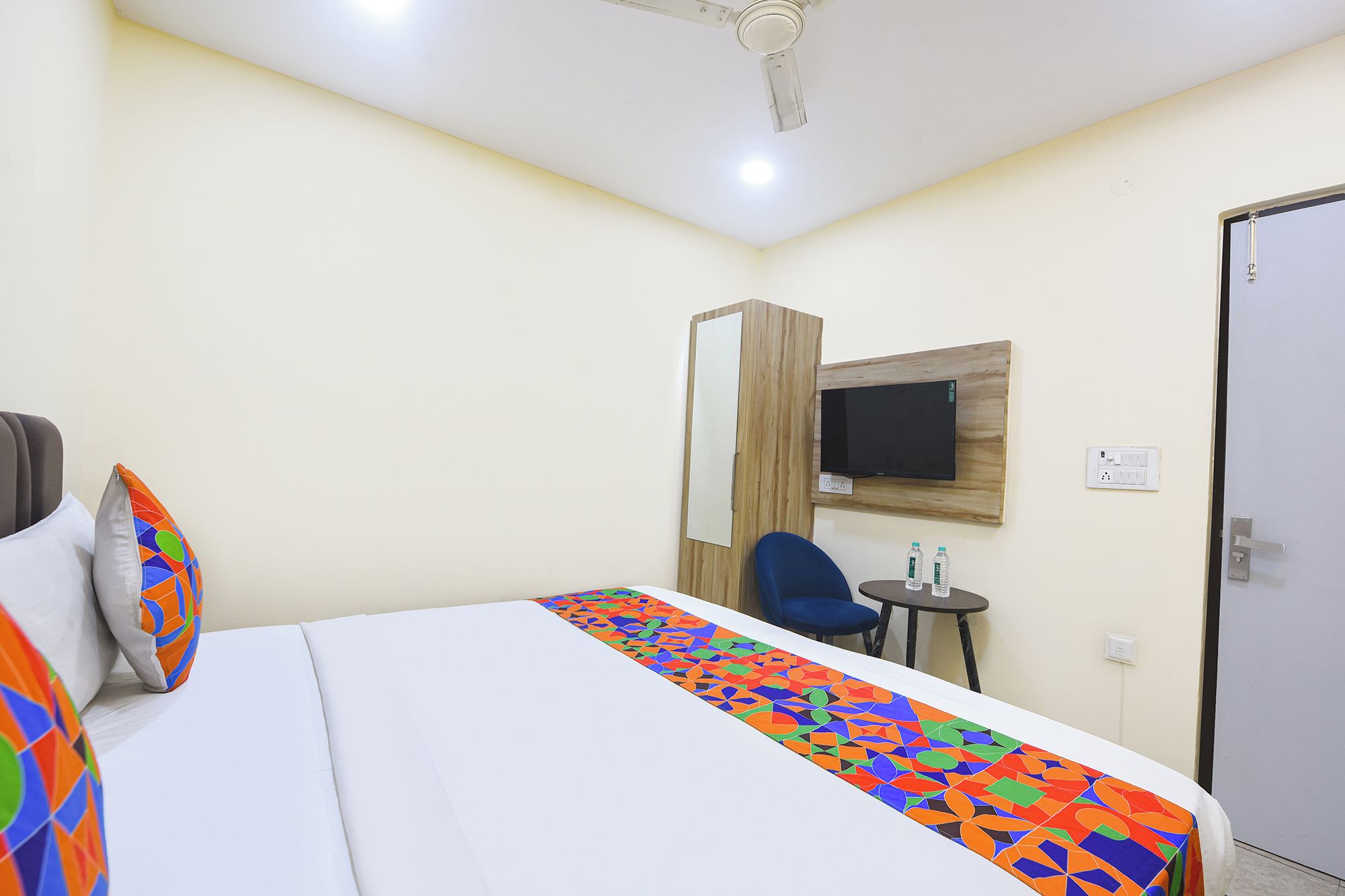 A hotel room with a bed, a television mounted on the wall, a small table with two bottles, and a door.