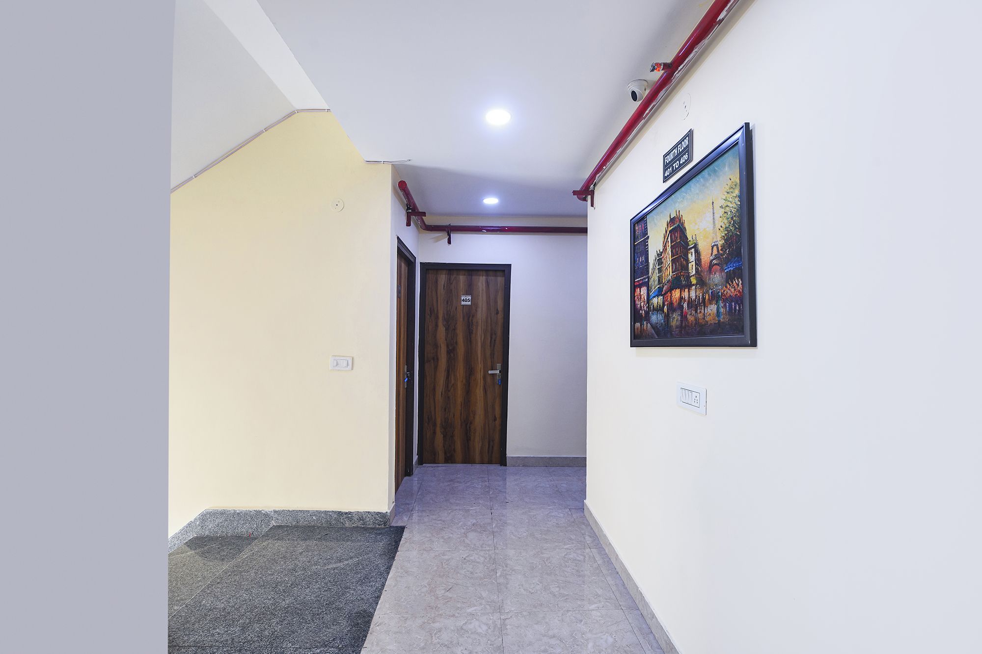 A hallway with a wooden door, a painting on the wall, and a red pipe running along the ceiling.