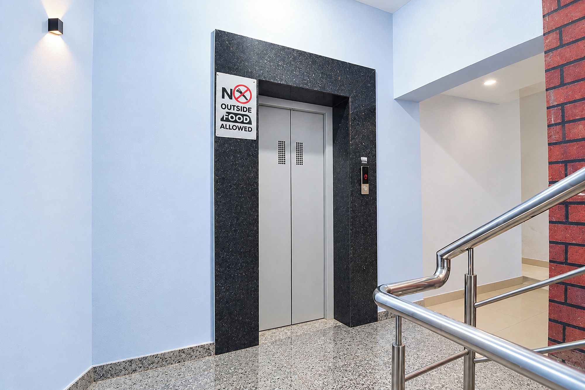 Un ascensor con un letrero que dice 'NO SE PERMITE COMIDA DE FUERA', una pantalla digital y un pasamanos de metal.