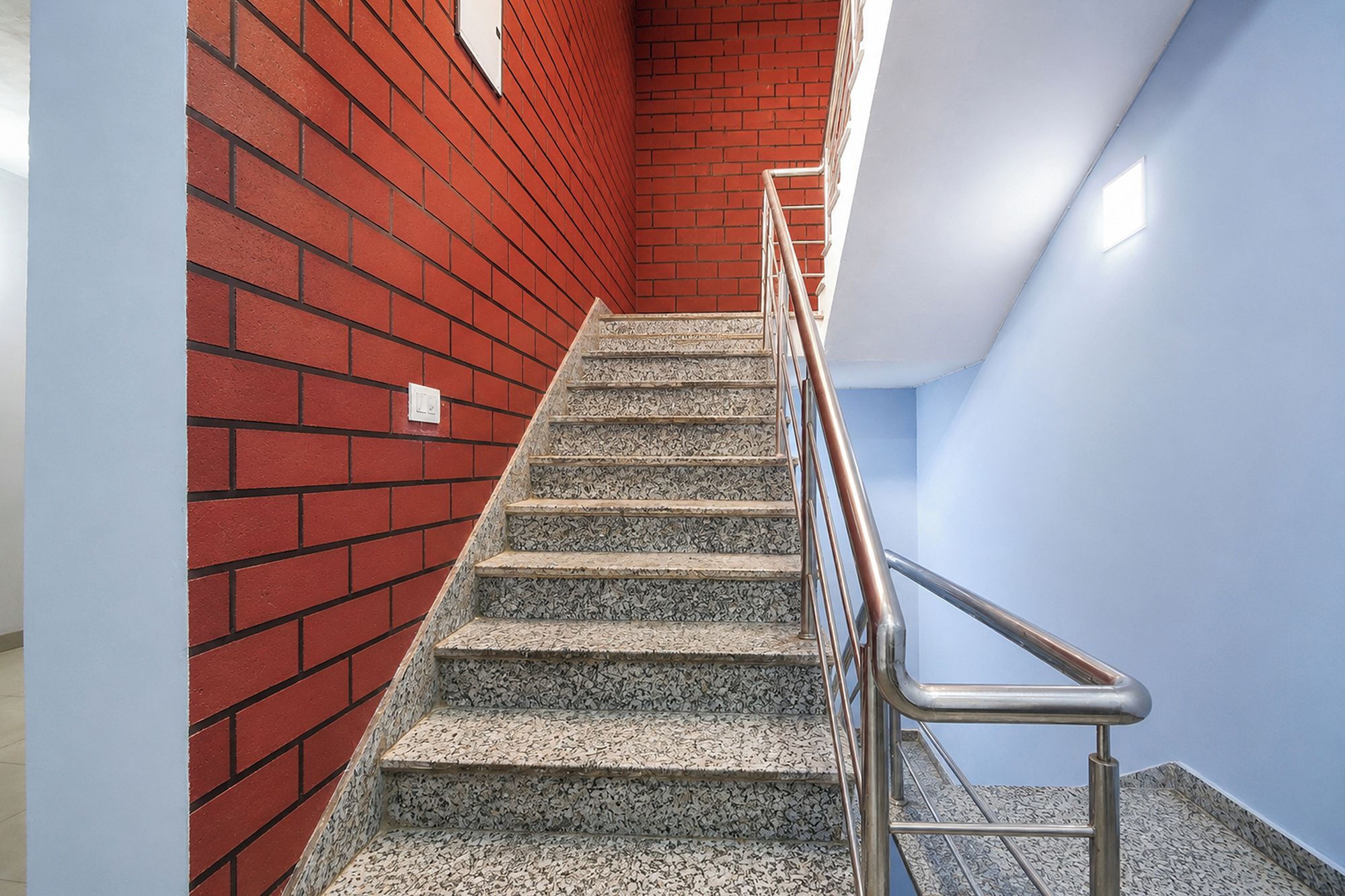 A staircase with a metal handrail, leading up to a landing with a red brick wall and a light blue wall.