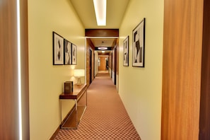 Corredor de hotel com quadros nas paredes, piso coberto por carpete e porta de madeira no lado direito.
