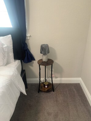A bedroom with a bed, a small table with a lamp, and a window with curtains. - Good Life! (Charlotte)
