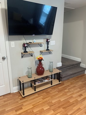 A living room with a flat-screen TV mounted on the wall, wooden shelves with decorative items, a vase with flowers, and a magazine on a wooden console table. - Good Life! (Charlotte)