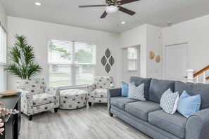 Un salon avec un canapé gris, des fauteuils à motifs, un ventilateur de plafond et une grande fenêtre avec des stores.