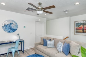 A living room with a ceiling fan, a wall-mounted artwork, a framed picture, a sofa with cushions, a desk with a chair, and a small plant. - Private Beach Access, Hot Tub, Balcony, Ocean Views, Brand New Furnishings, $300 in Free Tickets! (St. Augustine)